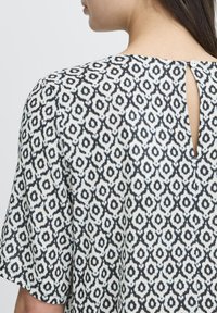 Blouse à manches courtes en tissu à motif noir et blanc avec une ouverture en forme de clé dans le dos et un détail de bouton. Présente un design floral géométrique.