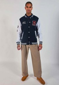 Veste varsity bleu marine et blanche avec des boutons, des poignets côtelés et un logo brodé, accompagnée de pantalons beiges et de mocassins bruns.