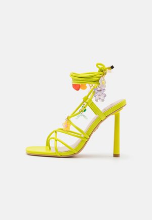 Żółte sandały na wysokim obcasie z wieloma paskami, ozdobione kolorowymi zawieszkami, w tym kwiatami i koralikami, o gładkim, błyszczącym wykończeniu.