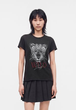 Jeune femme portant un t-shirt noir avec un visage de léopard et le texte "WILD Soul", associé à une jupe plissée noire, debout devant un fond blanc.