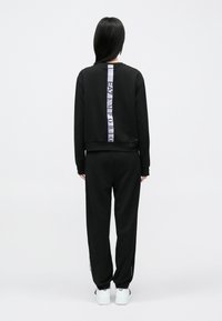 Osoba s dlhými tmavými vlasmi, oblečená v čiernej teplákovej súprave s vertikálnym pruhom "EA7 Emporio" na chrbte a bielymi teniskami, stojaca chrbtom.