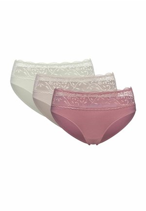 Woman with long dark hair wearing a beige lace bra and white lace-trimmed underwear standing against a plain light background.