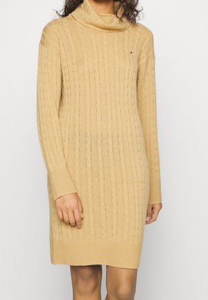 Femme portant une robe-pull beige à manches longues en maille torsadée avec un col bénitier sur un fond uni.
