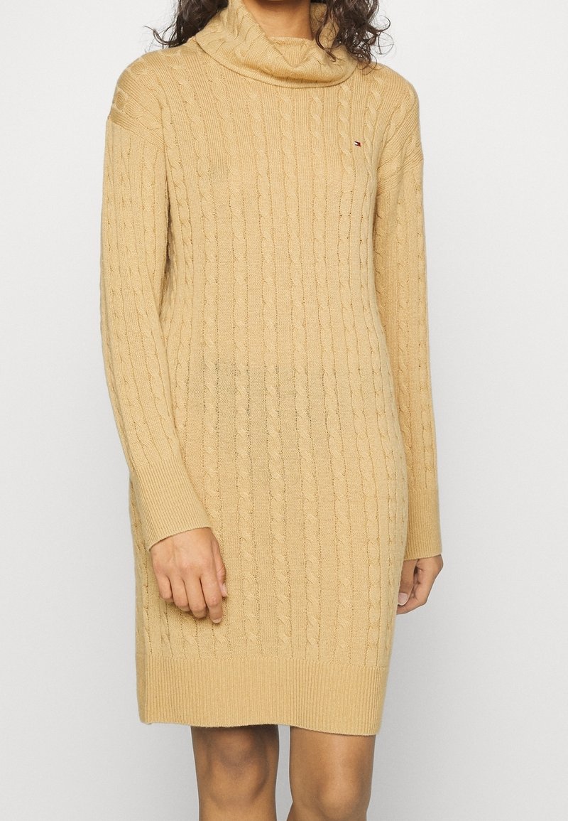 Femme portant une robe-pull beige à manches longues en maille torsadée avec un col bénitier sur un fond uni.