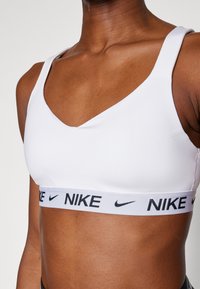 Hvit sports-bh med v-halsdesign og tykk svart elastisk bånd med "NIKE"-logo. Glatt stofftekstur og brede stropper.