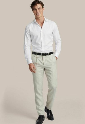 Man die een wit overhemd, lichtbeige broek met een zwarte riem en zwarte nette schoenen draagt, staand en glimlachend tegen een effen achtergrond.