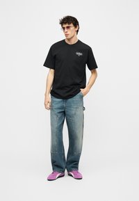 Νέος άνδρας που φοράει μαύρο μπλουζάκι με σχέδιο, ξεβαμμένα μπλε τζιν με λεπτομέρειες στις τσέπες, μωβ αθλητικά παπούτσια και γυαλιά ηλίου, στέκεται με το ένα χέρι στην τσέπη.