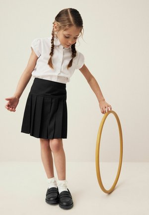 Witte kortekraag shirt met korte mouwen, zwarte geplooide rok, witte franje sokken en zwarte loafers. Meisje houdt een gele hoelahoep.
