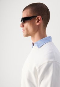 Black sunglasses with a glossy finish, rectangular shape, and tinted lenses, worn by a person in a white sweatshirt with a light blue collar.