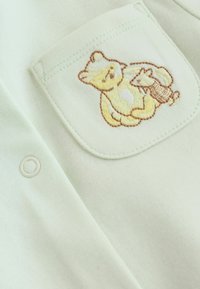 Tissu vert clair avec une petite poche présentant des personnages de dessins animés cousus, un ours et un écureuil jaunes avec un contour marron.