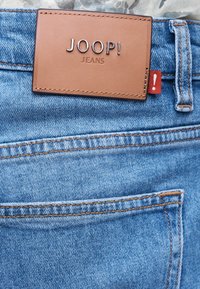 Καφέ δερμάτινο μπάλωμα με το λογότυπο "JOOP! JEANS" στην πίσω τσέπη από ανοιχτό μπλε τζιν με πορτοκαλί ραφές και κόκκινη ετικέτα που δείχνει ένα θαυμαστικό.