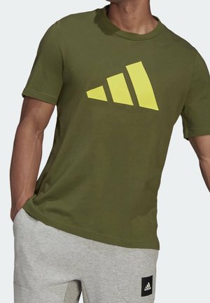 Mężczyzna w oliwkowozielonej koszulce z dużym żółtym logo Adidas oraz jasnoszarych spodniach dresowych z małą czarną łatką Adidas.