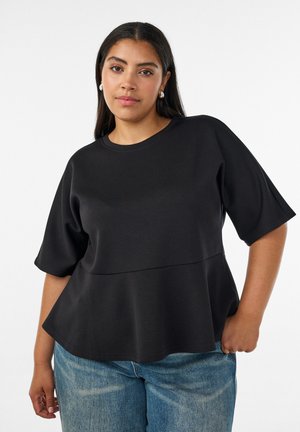 Ung kvinde med langt mørkt hår iført en sort kortærmet peplumtop og blå jeans mod en ensfarvet baggrund.