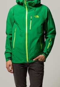 Grüne wasserdichte Jacke mit Kapuze, Reißverschluss vorne und zwei Brusttaschen. Mit gelben Reißverschlüssen und kontrastierenden schwarzen Akzenten. Leichtes Material.