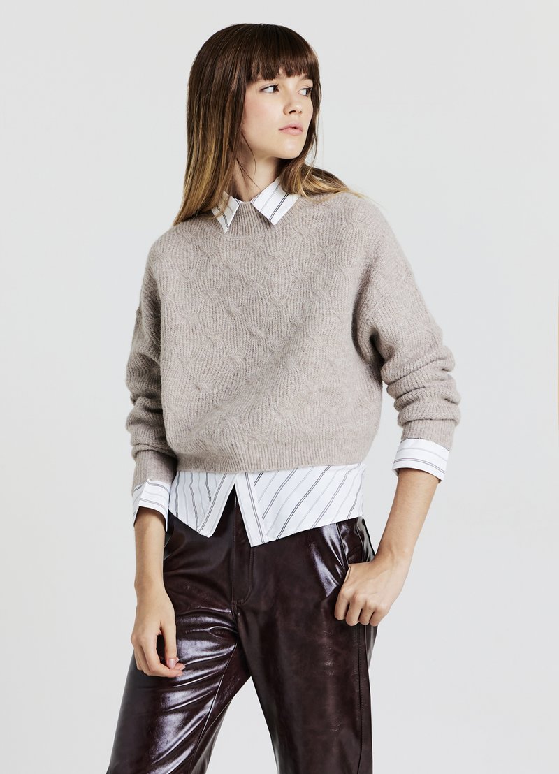 Maglione beige lavorato a maglia con un motivo testurizzato, indossato sopra una camicia colletto bianca e blu a righe, abbinato a pantaloni lucidi marroni scuri.