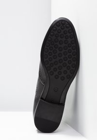 Musta kenkä, jossa on kuvioitu pohja pyöreillä kuvioilla, sileä yläosa rei'itettyine yksityiskohtineen ja pieni korko tukevuutta varten. Koko 37.