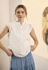 Femme aux cheveux blonds vêtue d'un chemisier blanc sans manches et d'une jupe bleue, debout les mains sur les hanches contre un mur beige.