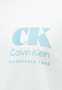 Λογότυπο Calvin Klein με μεγάλο στυλιζαρισμένο "CK" σε ανοιχτό μπλε, το όνομα της μάρκας από κάτω και το κείμενο "Ιδρύθηκε 1968" σε λευκό υφασμάτινο φόντο.