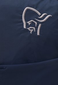 Tamsiai mėlynas audinys su siuvinėtu šviesiai pilku stilizuoto veido logotipu, akcentuojantis švarias linijas ir tekstūrinį paviršių.