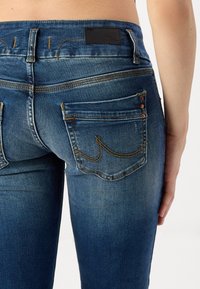 Vaqueros de mezclilla azules con costuras en contraste, diseño ajustado y bolsillos traseros visibles que presentan una etiqueta de marca y remaches de cobre.