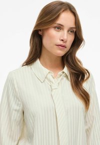 Jonge vrouw met lichtbruin haar, gekleed in een crèmekleurig gestreept shirt met een bijpassende stoffen knoopdetail op de kraag.
