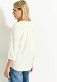 Blouse blanche et texturée avec un col rond, des poignets élastiques et un pli au dos, associée à un jean en denim bleu. Modèle vu de dos.