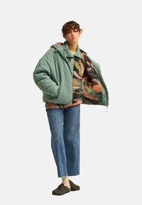 Young person wearing green quilted jacket open to show colorful patterned fleece, blue jeans, beige socks, and black slip-on shoes standing on white.
