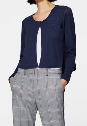 Femme portant un cardigan court bleu marine par-dessus un haut blanc et un pantalon taille haute gris à carreaux, les mains dans les poches.