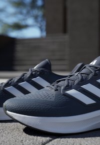 Baskets de sport gris foncé avec un tissu texturé, logo à trois rayures blanches et semelle blanche. Elles possèdent un col rembourré et des lacets.