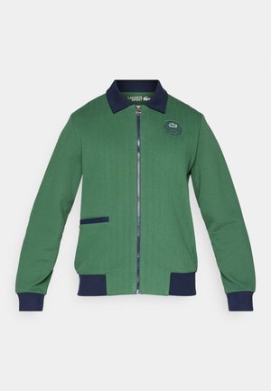 Giacca verde con zip, colletto e polsini blu notte. Tessuto a coste con una tasca frontale e logo ricamato sul petto.