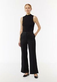 Zwarte geribbelde mouwloze top gecombineerd met high-waisted zwarte wijde broek. Model staat met de handen in de zakken en draagt platte zwarte schoenen.
