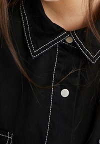 Gros plan sur une chemise noire à col avec des coutures blanches et des boutons-pression argentés, portée par une personne avec des mèches de cheveux bruns longs visibles.