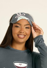 Szara maska do snu z nadrukiem przedstawiającym napój, noszona przez kobietę. Miękka tkanina z elastycznym paskiem. Pod spodem ciemna bluzka z długimi rękawami.