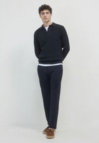 Pull noir en tricot côtelé avec un col, associé à un pantalon navy et des mocassins en daim marron. Le modèle se tient devant un fond clair.