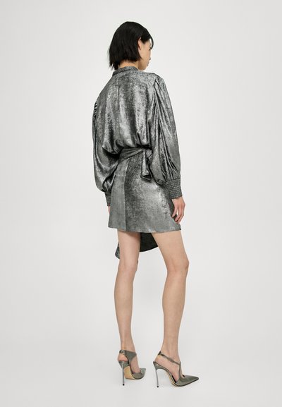 Metallisch zilveren jurk met opgeblazen mouwen en een bindtaille, voorzien van een korte asymmetrische zoom en textiele stof. Gecombineerd met grijze hoge hakken.