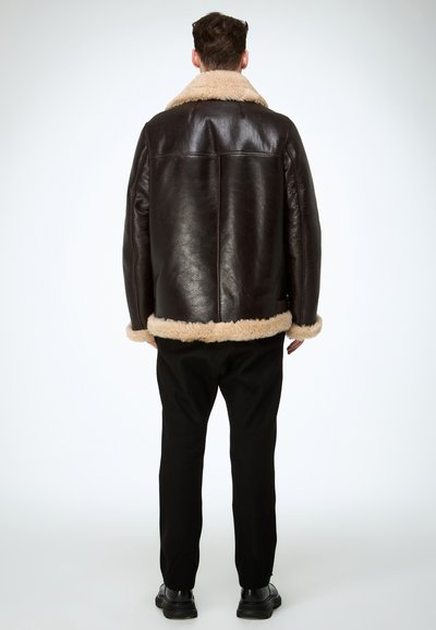 Veste en cuir marron avec une doublure et un col en peau lainée beige, présentant une coupe droite et un design minimaliste. Portée avec un pantalon noir.