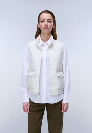 Donna che indossa un gilet trapuntato bianco sopra una camicia bianca con bottoni e pantaloni verde oliva, in piedi davanti a uno sfondo chiaro e uniforme.