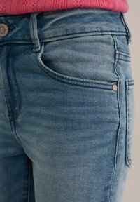 Světlé modré džínové kalhoty s mírným opotřebením, klasický design s pěti kapsami a kovovým zapínáním. Šité švy podél okrajů.