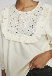 Blouse jaune clair à manches longues avec un col à volants et un détail en dentelle blanche complexe avec des motifs floraux. Tissu texturé avec une coupe décontractée.