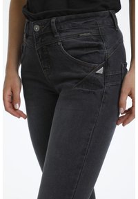 Jean en denim gris foncé avec une coupe slim, présentant des coutures contrastées, des poches avant et un accent logo métallique sur le côté.