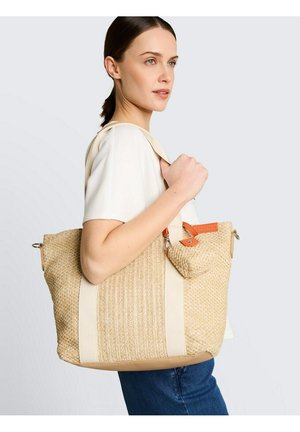 Vrouw in een wit shirt en een blauwe spijkerbroek draagt een grote beige geweven tas met oranje accenten over haar schouder.