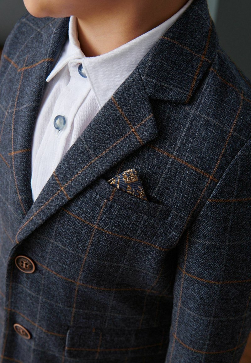 Enfant portant un blazer à carreaux foncés avec des lignes marron et une chemise blanche à col, montrant un mouchoir de poche à motif dans la poche du blazer.