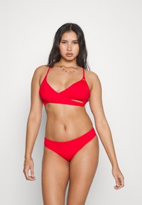 Rdeča bikini kombinacija, ki vključuje zgornji del s prečnim zapiranjem in tankimi naramnicami ter ujemajoče se nizke hlačke, izdelana iz gladkega, raztegljivega materiala.
