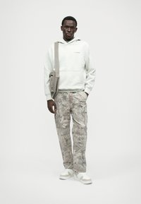 Szürke kapucnis pulóver elülső zsebbel, bézs katonai mintás cargo nadrággal, amely több zsebbel rendelkezik. A fehér sportcipők teszik teljessé az öltözetet.