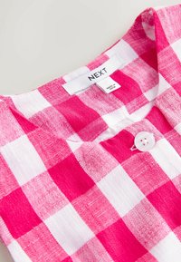Vêtement à carreaux roses et blancs avec un col rond, un bouton blanc et une étiquette de marque "NEXT" indiquant "Fabriqué en Inde".