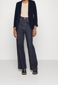 Blazer bleu marine, haut beige et pantalon large en denim foncé taille haute. Sandales noires à talons ouvertes complètent la tenue.