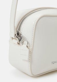 Bílá lakovaná krosbodi taška se stříbrným zipem, zaoblenými rohy a odnímatelným popruhem. Obsahuje malou visačku a decentní branding.