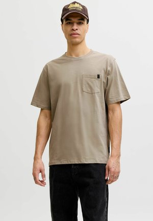 Fiatal férfi világosbarna rövid ujjú, mellzsebes pólóban és sötét nadrágban, egyszerű világos háttér előtt áll.