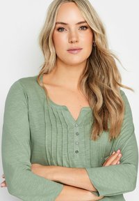 Grön, texturerad topp med V-ringning och tre knappar, med vertikala plisséer och trekvartslånga ärmar.