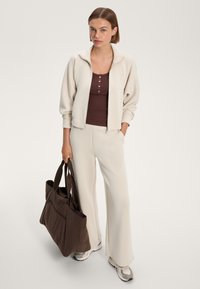 Svetlo bež jakna z rebrasto teksturo, v kombinaciji z rjavim topom in usklajenimi širokimi hlačami. V roki drži temno rjavo torbo.